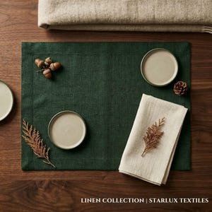 Op de afbeelding: Een donkergroene linnen placemat met twee kleine beige borden, een gevouwen beige linnen servet en decoratieve eikels en een dennenappel. De tekst "LINEN COLLECTION | STARLUX TEXTILES" staat onderaan.