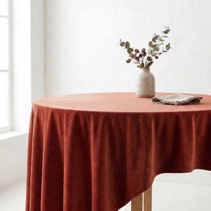 Könnte beinhalten: Ein runder, rostfarbener Samttischdecke auf einem Holztisch. Eine kleine Vase mit Grün und eine gefaltete Serviette liegen auf dem Tisch. Die Tischdecke hat ein weiches, luxuriöses Aussehen, geeignet für Mahlzeiten oder besondere Anlässe.