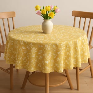May include: A round yellow tablecloth with a white floral pattern. The tablecloth is on a round wooden table with two wooden chairs. A vase with yellow and pink flowers is on the table.