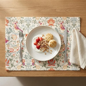 Pode incluir: Um lugar americano retangular com fundo creme e um padrão repetitivo de pássaros e motivos florais em laranja, rosa e azul. Um prato de cerâmica branca contém iogurte, morangos fatiados, granola e dois biscoitos, com um garfo e uma faca.