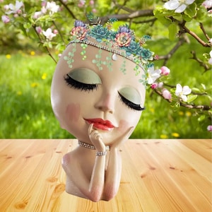 May include: A ceramic planter shaped like a woman's head with closed eyes and a crown of flowers and greenery. The planter is a light pink color with a white face and a pink blush on the cheeks. The planter is sitting on a wooden surface.