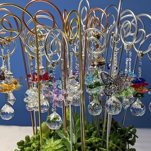 May include: A collection of crystal suncatchers with dragonfly, butterfly, and flower designs hanging from metal stands. The suncatchers are made of clear crystal with iridescent accents and are displayed against a blue background.