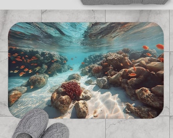 May include: A rectangular bath mat depicting an underwater scene with coral reefs, orange fish, and sunlight filtering through the water. The mat has a white sandy bottom and is surrounded by grey slippers and a grey towel.