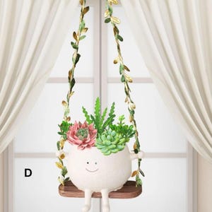 May include: A whimsical white ceramic planter shaped like a smiling character, holding a variety of succulents. The planter hangs from decorative vines with leaves. The planter is on a small wooden swing.