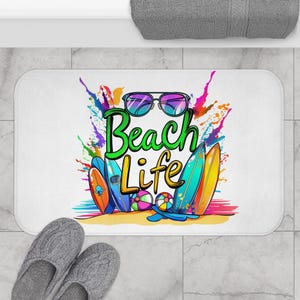 Może przedstawiać: Biała prostokątna mata łazienkowa z kolorowym wzorem "Beach Life". Wzór zawiera okulary przeciwsłoneczne, deski surfingowe i piłkę plażową. Tekst jest w kolorze zielonym i żółtym z kolorowymi plamami farby.