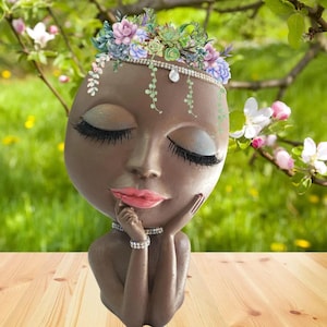 May include: A brown ceramic planter shaped like a woman's head with closed eyes and a flower crown. The planter has a delicate, feminine design and is perfect for displaying small plants.