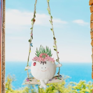 Op de afbeelding: Een grillige witte plantenbak in de vorm van een lachend karakter, hangend aan een schommel. De plantenbak is gevuld met vetplanten en roze bloemen. De schommelkoorden zijn versierd met bladeren en kleine bloemen.