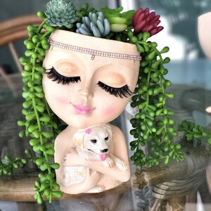 May include: A decorative planter shaped like a woman's face, with long eyelashes, pink cheeks, and a rhinestone headband. The planter holds a small white dog and is adorned with faux succulents and trailing greenery.