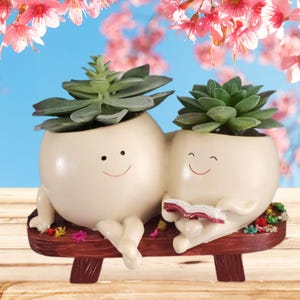 May include: Two white ceramic planters shaped like people, each with a smiling face, sitting on a wooden bench. The planters are holding succulent plants and the one on the right is holding a red book.