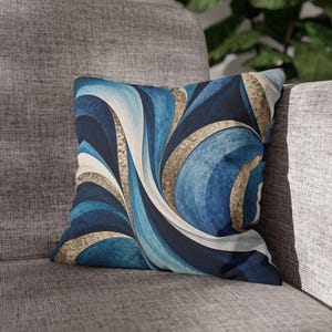 Puede incluir: Un cojín cuadrado con un patrón abstracto dinámico de ondas arremolinadas en tonos azul marino, azul claro y blanco, acentuado con bandas texturizadas de tono dorado que siguen las líneas curvas por toda la superficie de la tela.