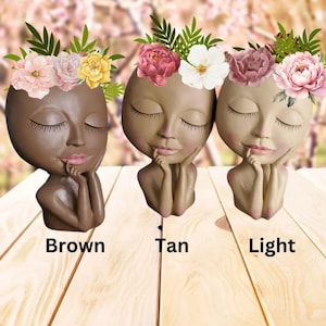 May include: Three ceramic planter pots shaped like faces with closed eyes and a flower crown. The pots are in three different colors: brown, tan, and light. The text below each pot reads "Brown", "Tan", and "Light".