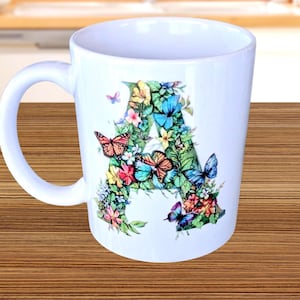 Może przedstawiać: Biały ceramiczny kubek z kwiatowym i motylkowym wzorem. Litera "A" jest utworzona z zielonych liści, kwiatów i motyli w odcieniach pomarańczu, błękitu, różu i żółci. Kubek ma zakrzywiony uchwyt.