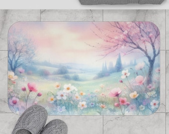 May include: A rectangular bath mat with a watercolour landscape design. The mat depicts a field of pink, white, and blue flowers, trees, and a pastel sky. Grey slippers are in the foreground. The mat is likely 70 cm x 50 cm.
