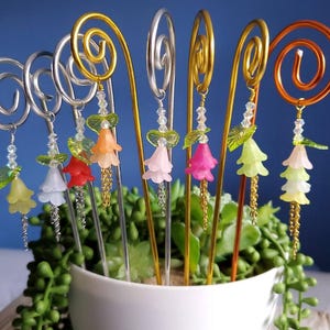 Puede incluir: Una colección de accesorios para el cabello florales decorativos. Cada accesorio para el cabello presenta un diseño de espiral o remolino de alambre en plata, oro o cobre. Los accesorios están adornados con coloridos dijes de flores, cuentas y detalles de hojas, y se exhiben en una maceta blanca.