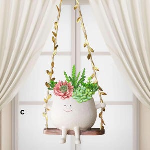 Puede incluir: Una maceta de cerámica blanca caprichosa con forma de personaje, suspendida de un columpio dorado. La maceta está llena de una variedad de suculentas artificiales de colores. El columpio cuelga frente a una ventana con cortinas blancas.