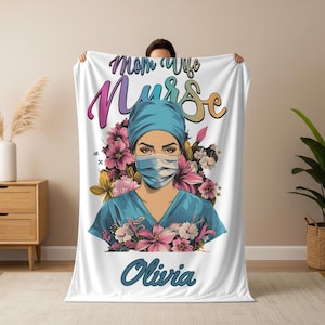 Puede incluir: Manta blanca con una ilustración de enfermera y el texto "Mom Wife Nurse Olivia". La enfermera lleva uniforme azul, gorro y mascarilla, rodeada de motivos florales rosas, morados y blancos.