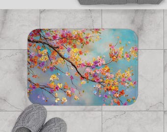 May include: Rectangular bath mat featuring a vibrant floral design. The artwork depicts a tree branch laden with colorful blossoms in shades of pink, yellow, orange, and purple against a light blue background. The mat is placed on a tiled floor.