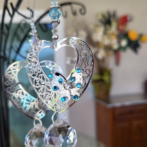 May include: A decorative heart-shaped suncatcher with intricate cut-out patterns and a butterfly design, adorned with blue crystals. Two clear crystal spheres hang below. The suncatcher is suspended by a string with blue beads.