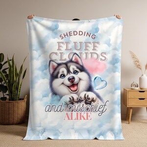 May include: A soft, light blue blanket featuring a cartoon husky dog with a smiling face, surrounded by clouds. The blanket has the text "Shedding Fluff Clouds and Mischief Alike" in a whimsical font. A pink cloud and a blue heart are also featured.