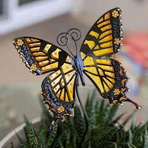 Può includere: Una farfalla decorativa con ali nere e gialle, dettagliata con piccoli accenti dorati. La farfalla ha un corpo in metallo nero ed è posizionata sopra una pianta verde. Le ali sono aperte, mostrando il design intricato.