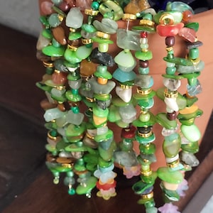 Puede incluir: Colección de joyas con cuentas con una variedad de piedras verdes, marrones y doradas. Las cuentas están ensartadas con detalles dorados. Las joyas se exhiben sobre un fondo marrón.