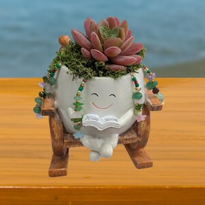 May include: A whimsical white ceramic planter shaped like a smiling character, holding a book and seated in a wooden chair. The planter is filled with a succulent plant and decorative beads. The background is a blurred view of the ocean.