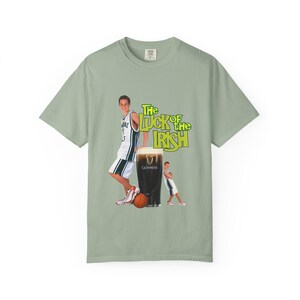 May include: Sage green t-shirt featuring a graphic of a basketball player leaning on a pint of Guinness, with a basketball and the text "The Luck of the Irish" in green and yellow. The Guinness glass has a white head.