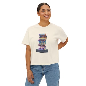 May include: Cream-colored t-shirt featuring a stack of colorful VHS tapes on a black rectangular base. The tapes have various titles and colors, evoking a retro aesthetic. The shirt is a relaxed fit.