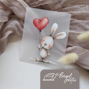 Könnte beinhalten: Ein rechteckiges Bügelbild mit einem hellbraunen Cartoon-Hasen mit langen Ohren und rosa Wangen, der einen roten herzförmigen Luftballon hält. Das Design enthält den Text Bügelmotiv sowie ein kleines Bügeleisen-Symbol in der unteren Ecke.