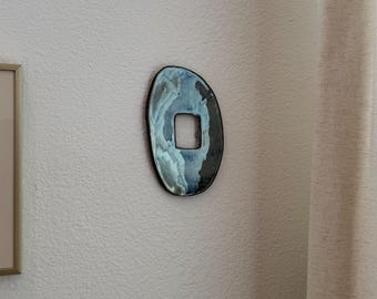 Può includere: Una placca di copertura per interruttore della luce in ceramica di forma ovale, caratterizzata da una smaltatura lucida con motivi vorticosi in blu navy, azzurro e bianco. La placca presenta un bordo organico irregolare e un foro centrale.