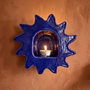 May include: A deep blue ceramic wall-mounted candle holder shaped like a stylized sun with jagged rays. The center features a recessed arched niche containing a small, lit white tea light candle with a visible flame, creating a warm glow.