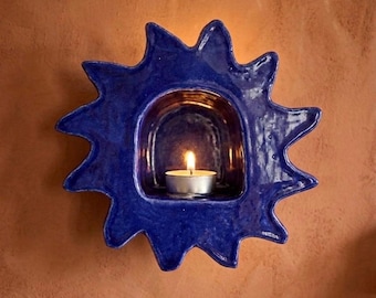 Può includere: Un portacandele da parete in ceramica blu intenso a forma di sole stilizzato con raggi frastagliati. Il centro presenta una nicchia ad arco incassata contenente una piccola candela tea light bianca accesa con fiamma visibile.