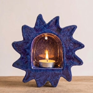 May include: A sun-shaped ceramic candle holder with a deep blue, mottled glaze finish. The center features an arched alcove containing a small, lit tea light candle. The outer edge has pointed rays extending outward in a starburst pattern.