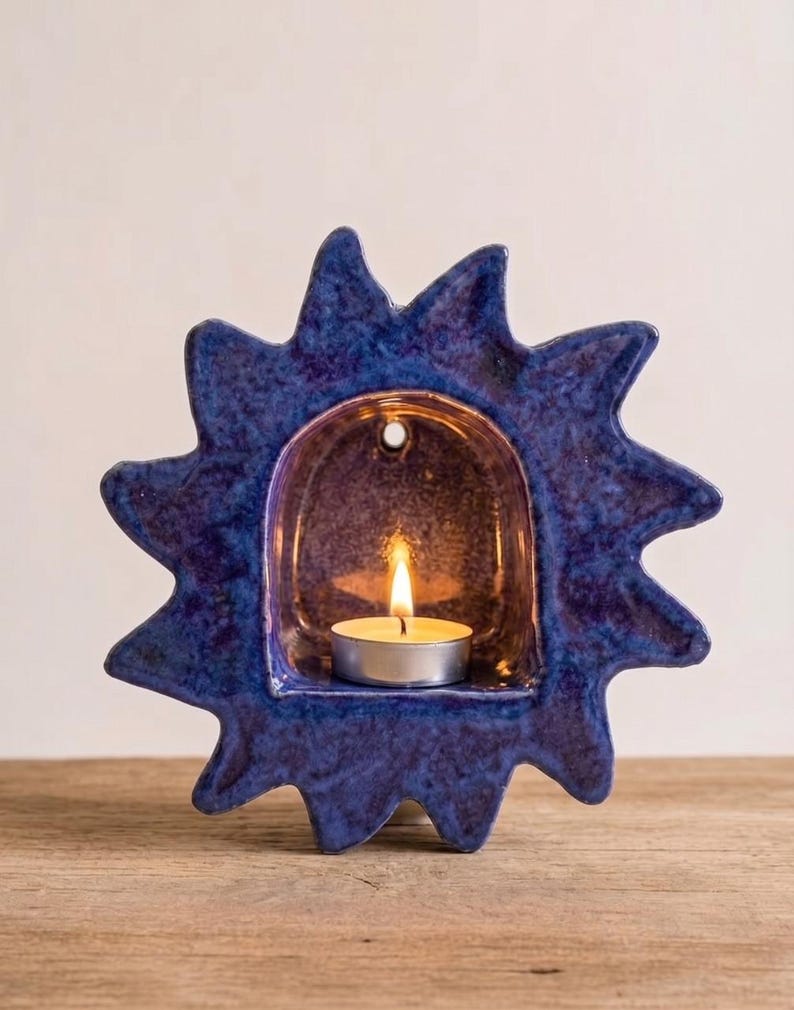 May include: A sun-shaped ceramic candle holder with a deep blue, mottled glaze finish. The center features an arched alcove containing a small, lit tea light candle. The outer edge has pointed rays extending outward in a starburst pattern.