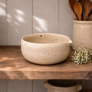 Puede incluir: Un cuenco de cerámica moteada de color blanquecino con un pico vertedor. El cuenco está sobre una estantería de madera, con cucharas de madera en un tarro de cerámica a juego en el fondo. Una ramita de flores blancas añade un toque delicado.