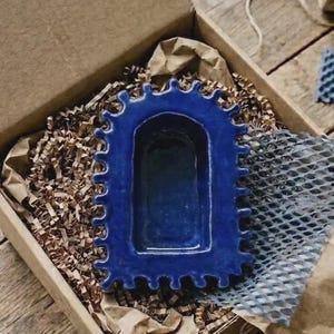 Pu&ograve; includere: Un vassoio in ceramica blu profondo con un centro rettangolare e un bordo smerlato e ondulato. L'oggetto presenta una finitura lucida e liscia con una conca incassata, presentato in una scatola di cartone con riempitivo di carta marrone.