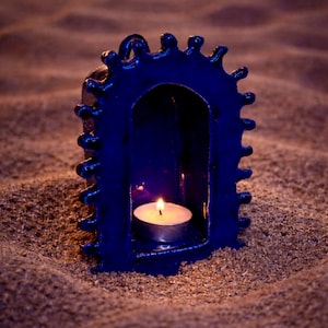 Puede incluir: Un portavelas azul oscuro con una vela encendida en su interior. El portavelas tiene una abertura arqueada y un diseño dentado en la parte superior. Se encuentra sobre una superficie arenosa.