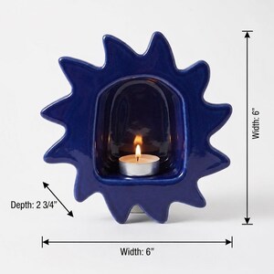 May include: A dark blue ceramic candle holder shaped like a sun with a central alcove containing a lit tea light candle. The object measures 6 inches in width and 6 inches in height, with a depth of 2 3/4 inches as indicated by labels.