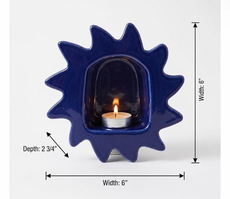 May include: A dark blue ceramic candle holder shaped like a sun with a central alcove containing a lit tea light candle. The object measures 6 inches in width and 6 inches in height, with a depth of 2 3/4 inches as indicated by labels.