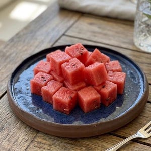 May include: A pile of bright red, cubed watermelon pieces with small black seeds arranged on a round, dark blue ceramic plate with a thick, textured brown rim. The plate is positioned on a wooden surface next to a silver metal fork.