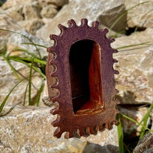 Pu&ograve; includere: Una scultura in ceramica caratterizzata da un esterno testurizzato marrone-rossastro scuro con un perimetro frastagliato a forma di ingranaggio. Il centro presenta un'apertura ad arco profonda e liscia con una finitura interna rosso-arancio.