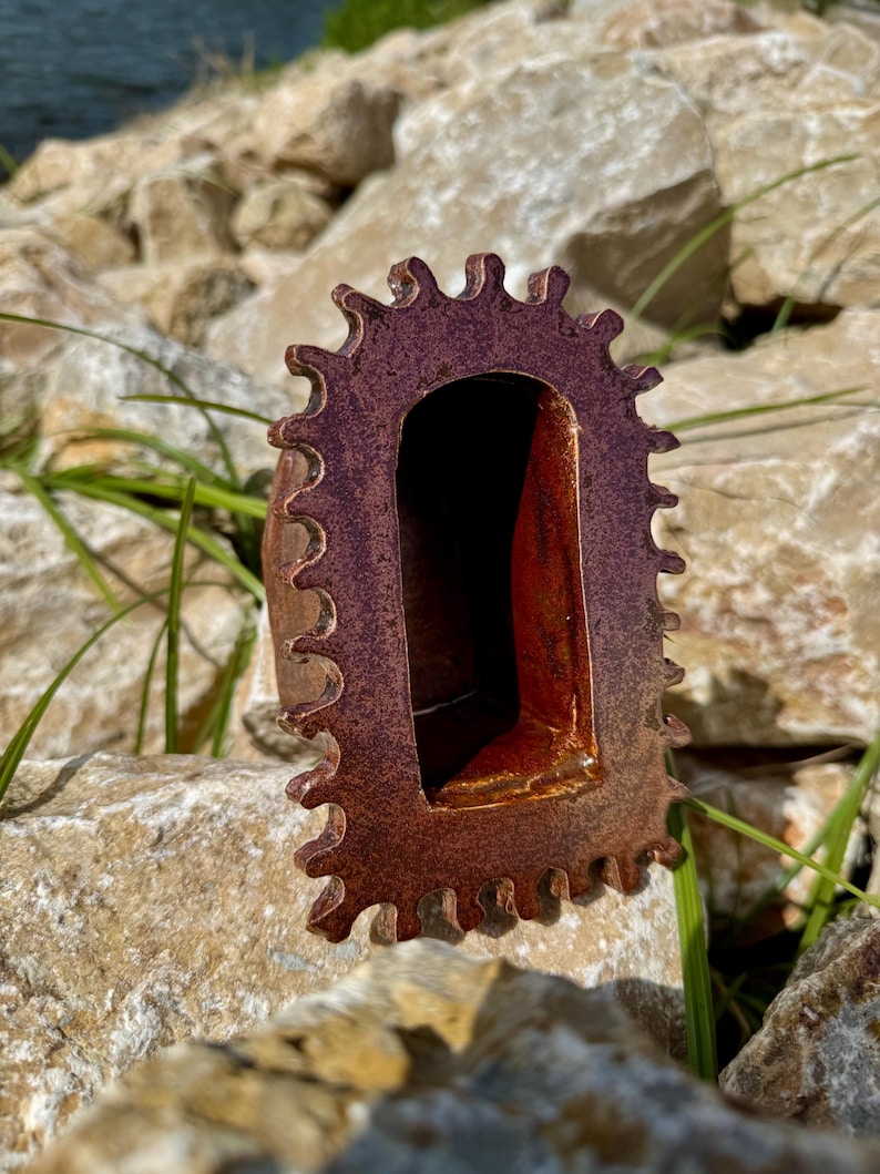 Pu&ograve; includere: Una scultura in ceramica caratterizzata da un esterno testurizzato marrone-rossastro scuro con un perimetro frastagliato a forma di ingranaggio. Il centro presenta un'apertura ad arco profonda e liscia con una finitura interna rosso-arancio.