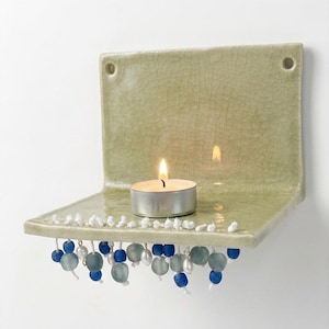 May include: A sage green ceramic wall shelf with a lit tealight candle. The shelf is decorated with white, blue, and pearl-like beads hanging from the bottom edge. The shelf has a crackle glaze finish and two holes for hanging.