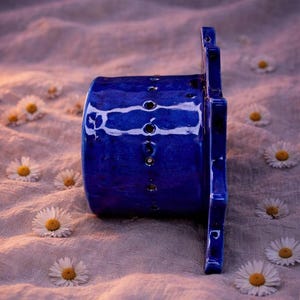 May include: A cylindrical cobalt blue ceramic object with a glossy finish and a vertical row of small circular perforations along its side. The object features a flat, star-shaped extension on one end with additional cutouts and edges.
