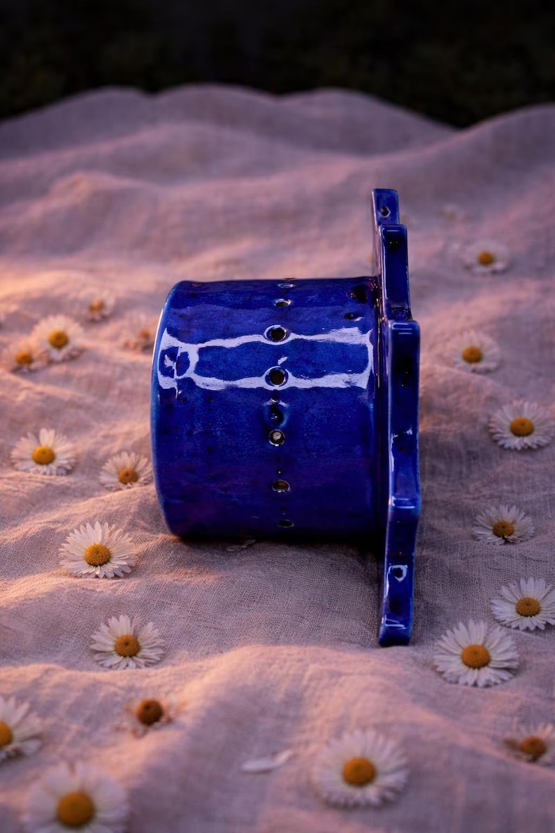 May include: A cylindrical cobalt blue ceramic object with a glossy finish and a vertical row of small circular perforations along its side. The object features a flat, star-shaped extension on one end with additional cutouts and edges.