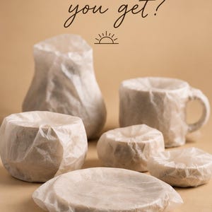 May include: A collection of various ceramic objects wrapped in translucent white tissue paper. The text "what will you get?" appears at the top with a small sun icon, and "A SURPRISE PIECE FROM MY STUDIO" is printed at the bottom.