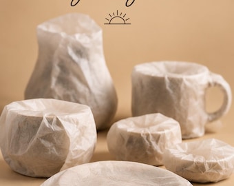Può includere: Una collezione di vari oggetti in ceramica avvolti in carta velina bianca traslucida. Il testo "what will you get?" appare in alto con una piccola icona del sole, e "A SURPRISE PIECE FROM MY STUDIO" è stampato in basso.