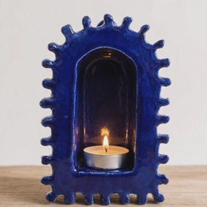 Pu&ograve; includere: Un portacandele in ceramica blu intenso con finitura lucida e un bordo esterno smerlato e irregolare. La struttura presenta una nicchia centrale ad arco contenente una piccola candela scaldavivande argentata con una fiamma accesa.