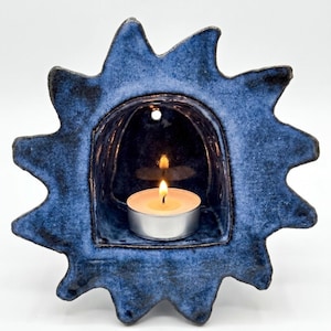 May include: A sun-shaped ceramic candle holder with a deep blue, mottled glaze. The center features an arched alcove containing a small, round tea light candle with a lit flame. A small circular hole is visible at the top of the alcove.
