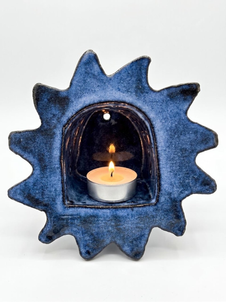 May include: A sun-shaped ceramic candle holder with a deep blue, mottled glaze. The center features an arched alcove containing a small, round tea light candle with a lit flame. A small circular hole is visible at the top of the alcove.